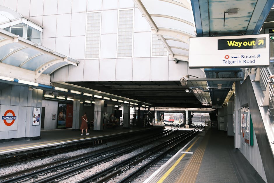 Photograph of the exterior exit area of Earls Court station, showing the underground platform entrance under a curved, transparent roof. Inside the station, a few commuters are visible walking along the platform, which is lined with safety yellow and white markings. The station's signage includes a black and yellow 'Way out' sign with an arrow pointing to the right, and a blue and white sign indicating bus services to Talgarth Road. Adjacent to the station entrance, there are large advertisements and a map poster on the white tiled walls. The scene captures the modern, urban environment typical of London transport hubs, ready to facilitate home relocation or furniture transport logistics connected to removals service providers like Man and Van Earls Court, with a focus on efficient passenger movement and accessible exit pathways.