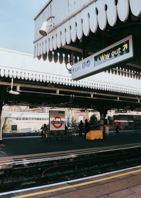 Outside Earls Court station, the image shows a platform area with black railway tracks in the foreground, accompanied by a yellow safety line. The station’s white canopy with scalloped edges extends overhead, providing shelter for waiting passengers. A prominent green and white 'Way out' sign hangs from the ceiling, indicating the exit direction. Beneath the canopy, there are several people, some sitting on wooden benches, and others standing near the platform edge. To the left, a large advertisement for Golders Green is visible on a wall behind the seated individuals. The scene includes elements of urban infrastructure, such as metal railings, a yellow tactile paving strip along the platform, and various structural supports, all illuminated by natural daylight. Man and Van Earls Court occasionally handle the logistics of furniture transport and packing during home relocation, often involving careful loading and unloading processes at stations like this, adhering to safety and access tips for efficient moving operations.