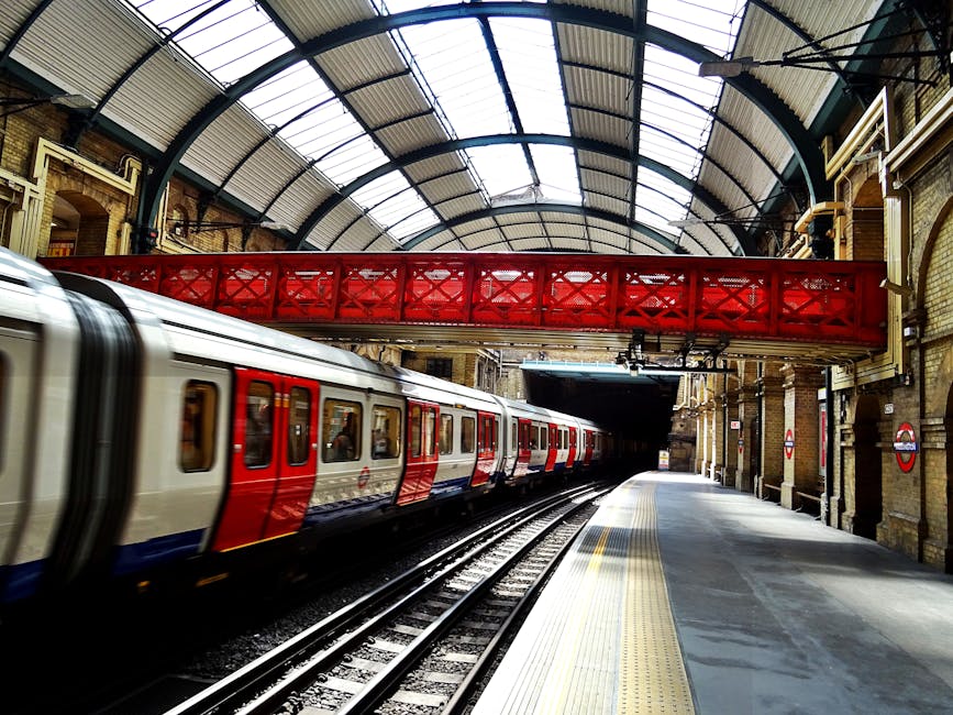 Photograph of the exterior exit area of Earls Court station, showing the underground platform entrance under a curved, transparent roof. Inside the station, a few commuters are visible walking along the platform, which is lined with safety yellow and white markings. The station's signage includes a black and yellow 'Way out' sign with an arrow pointing to the right, and a blue and white sign indicating bus services to Talgarth Road. Adjacent to the station entrance, there are large advertisements and a map poster on the white tiled walls. The scene captures the modern, urban environment typical of London transport hubs, ready to facilitate home relocation or furniture transport logistics connected to removals service providers like Man and Van Earls Court, with a focus on efficient passenger movement and accessible exit pathways.