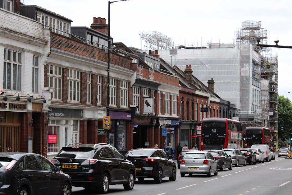 A street scene on Earls Court Road showing a line of parked vehicles including black, silver, and white cars and two red double-decker buses, with a row of traditional brick and white-painted terraced houses along the pavement. Some of the buildings have large windows and storefronts, with signage visible. In the background, scaffolding and construction netting cover a building under renovation. The pavement is occupied by pedestrians, and the area appears busy with traffic and moving activity. The sky is overcast, providing diffuse lighting across the scene, and the overall environment reflects a typical urban residential and commercial area suitable for professional removals and home relocation services, with visible loading and unloading activities associated with urban house moving processes. Man and Van Earls Court operates in this context, providing furniture transport and packing and moving services.